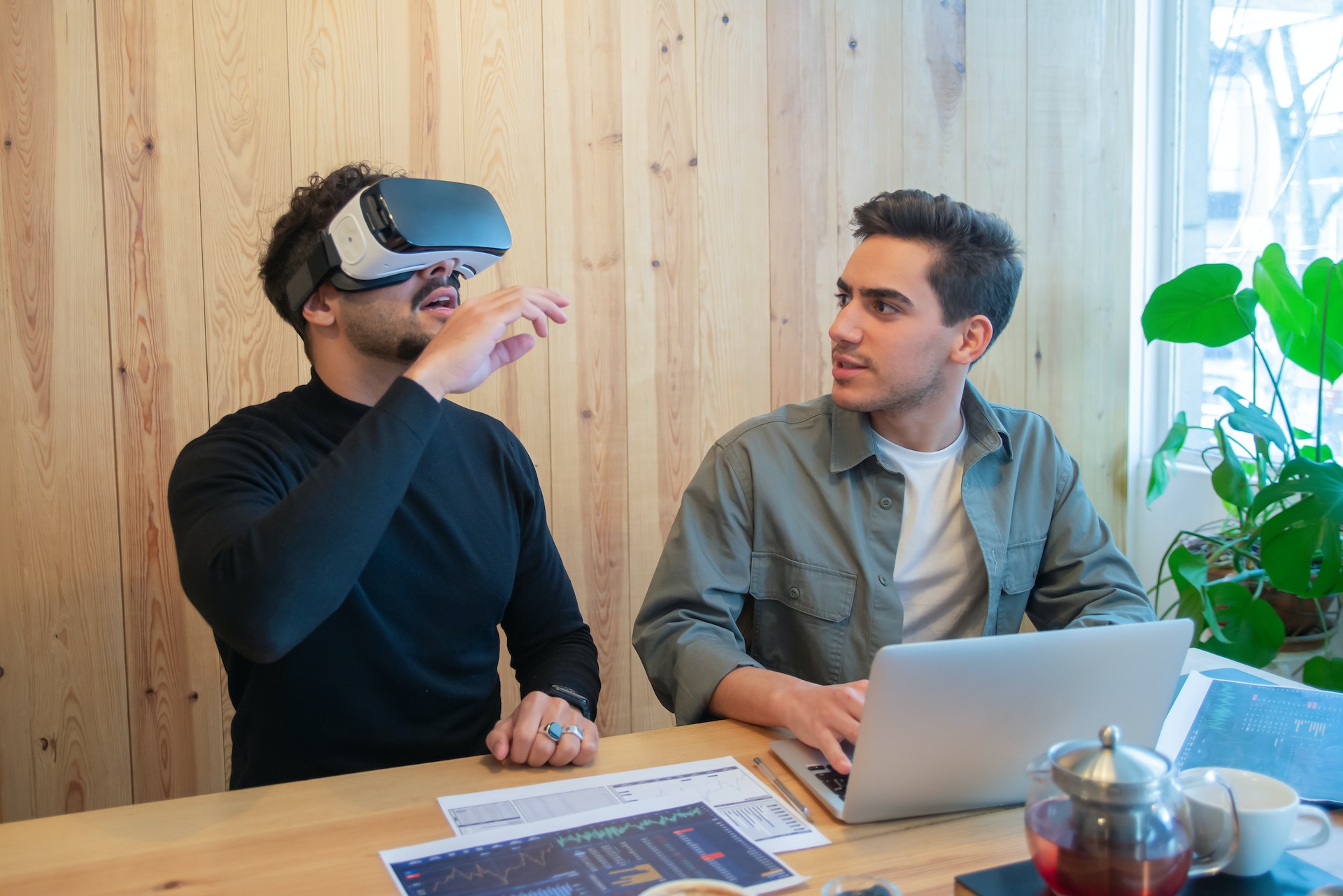 Two people sitting together at a desk. One is using a VR headset.